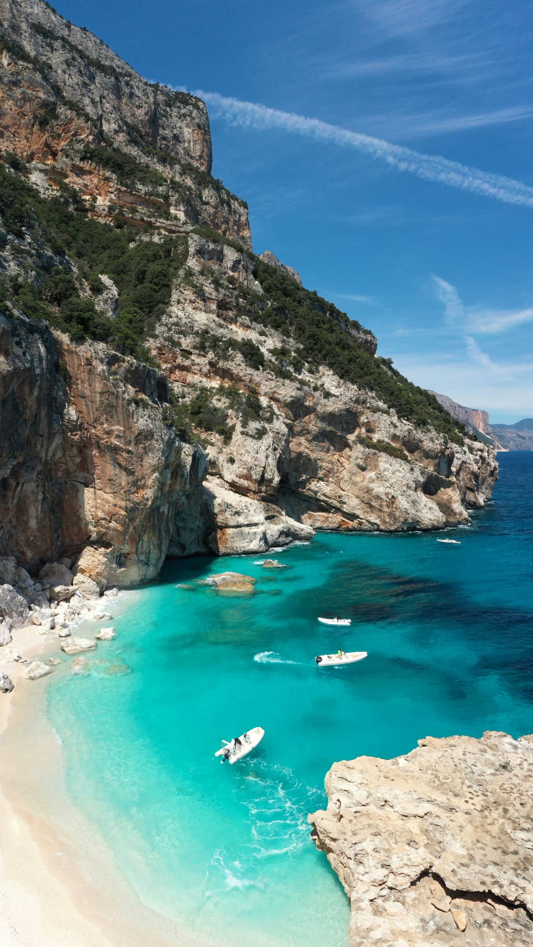 Small boats anchored in a turquoise cove beneath limestone cliffs on the Sardinian coast